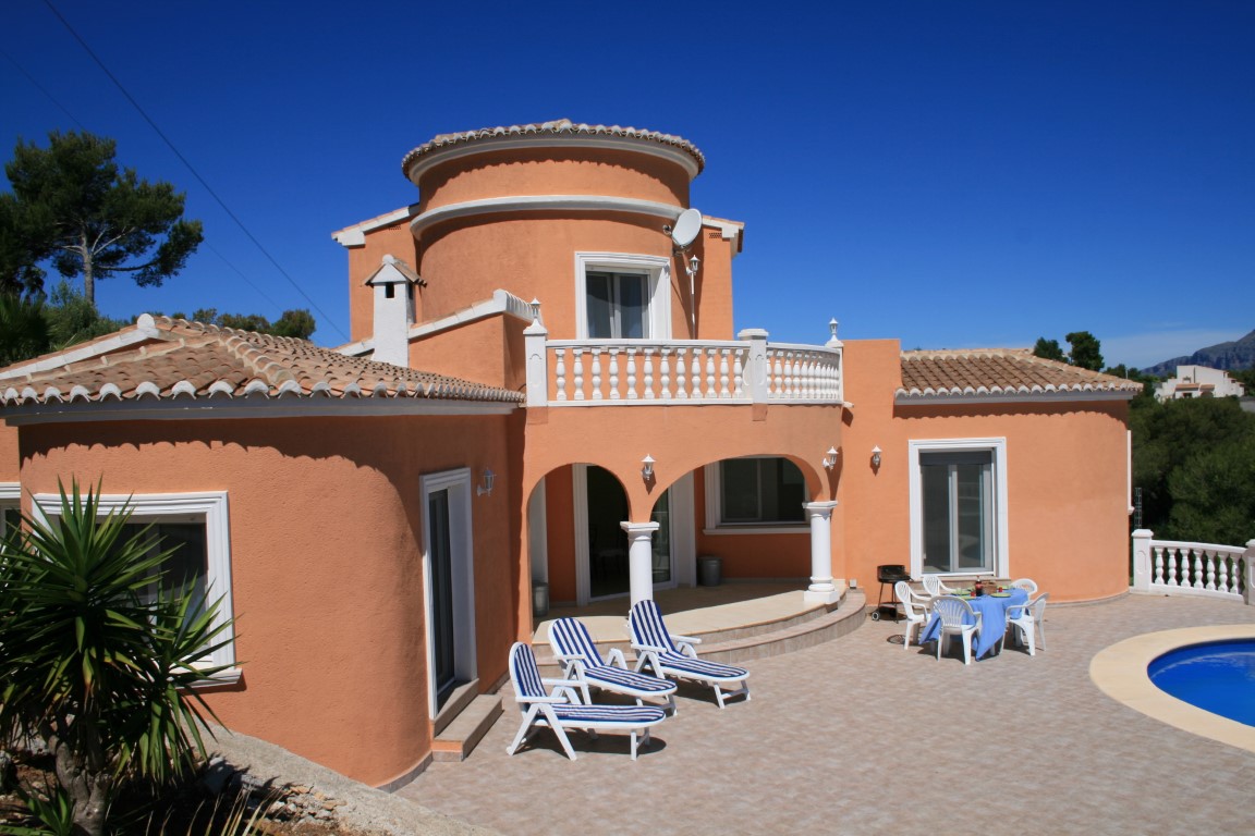 Extérieur d'une villa terre cuite avec tour ronde, porche voûté et grande terrasse carrelée équipée de bains de soleil près de la piscine.