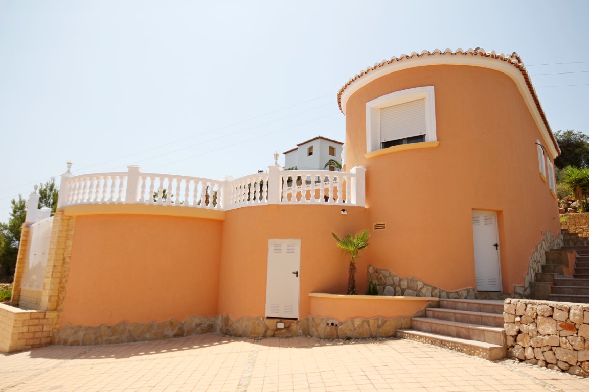 Façade extérieure courbe aux tons terre cuite avec balustrades blanches, soubassement en pierre et terrasse pavée sous un ciel dégagé.