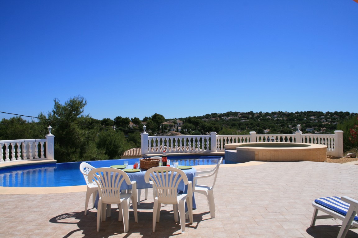 Grande terrasse extérieure avec piscine incurvée, jacuzzi circulaire intégré et espace dînatoire offrant une vue panoramique sur les collines de Javea.