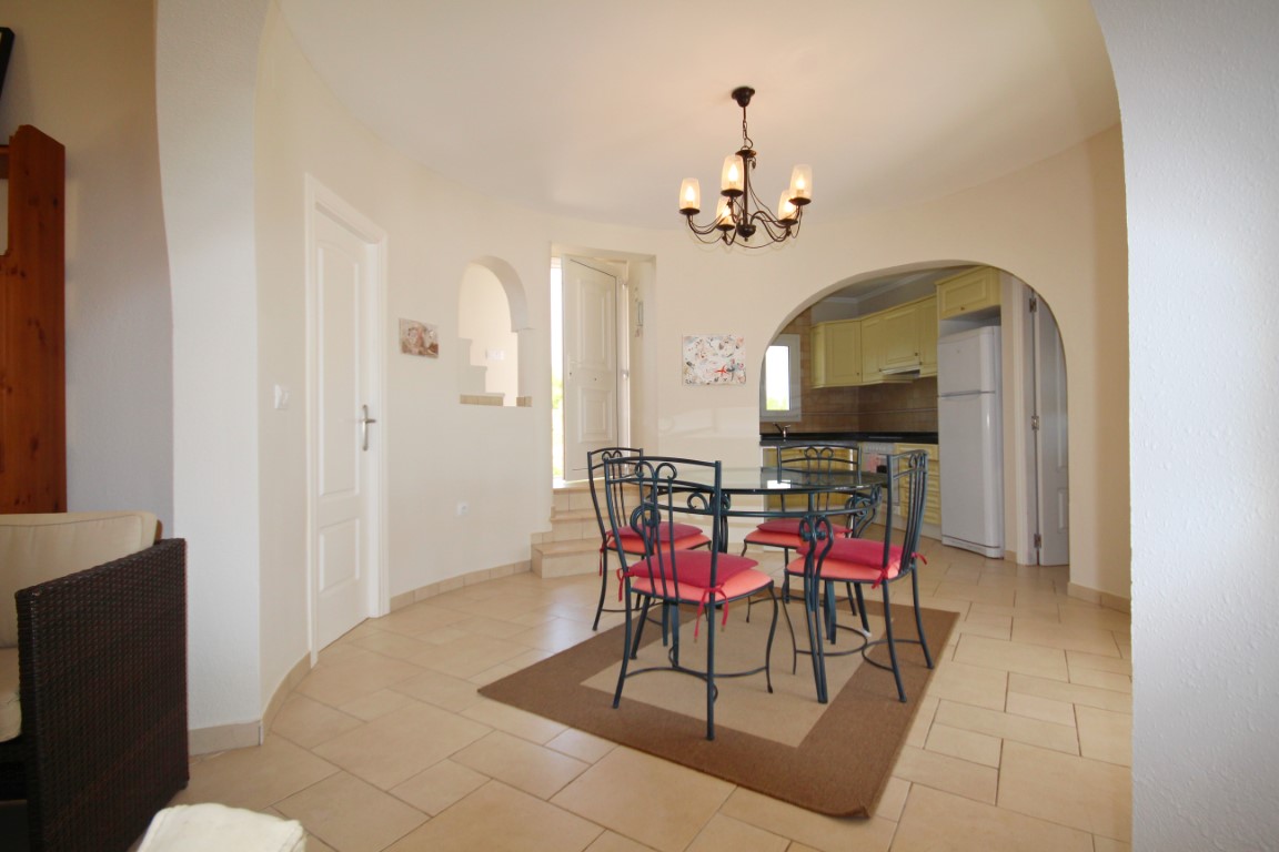 Salle à manger avec table en verre et chaises en fer forgé, ouverte par une arche sur une cuisine aménagée aux tons clairs et électroménager blanc.