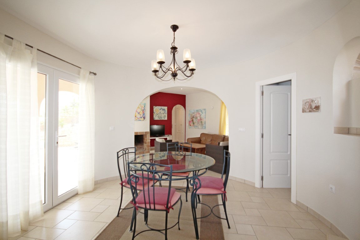Salle à manger lumineuse avec table en verre et chaises en fer forgé, ouverte sur le salon par une arche élégante révélant une cheminée.