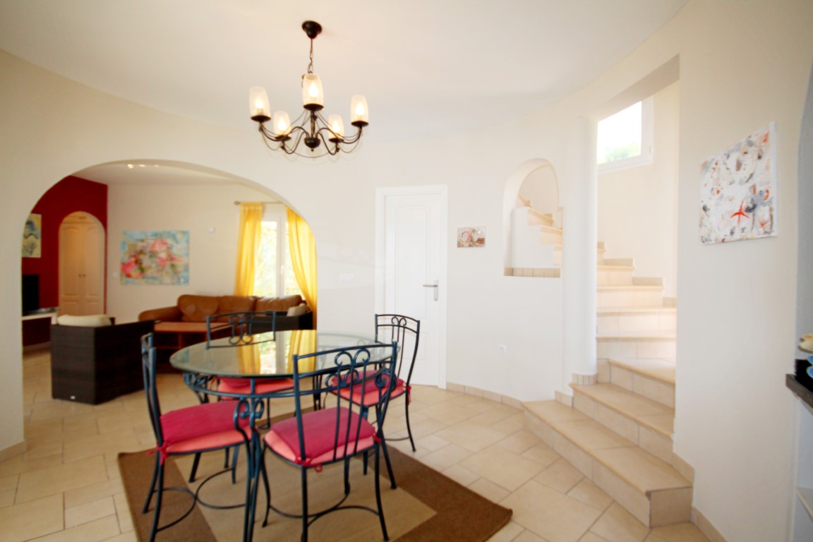 Salle à manger lumineuse avec table en verre et chaises en fer forgé, s'ouvrant sur le salon par une arche et jouxtant l'escalier intérieur carrelé.