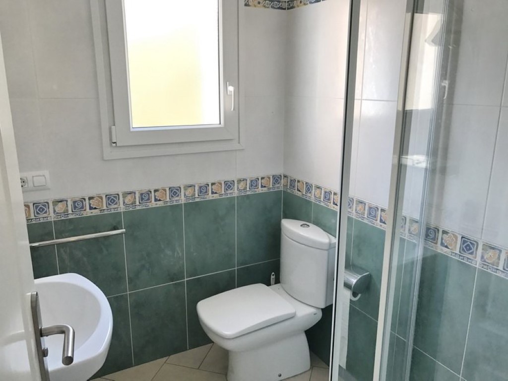 Salle de bain avec carrelage mural vert et blanc, toilettes en porcelaine, lavabo et douche avec paroi vitrée. Une fenêtre assure une luminosité naturelle.