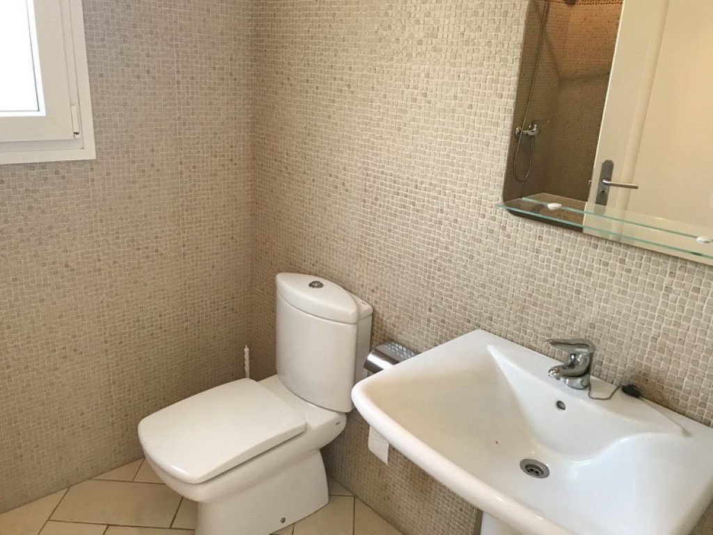 Salle de bain avec murs en mosaïque beige, lavabo en céramique blanche et miroir avec tablette en verre. Comprend des carreaux de sol neutres et une fenêtre latérale.