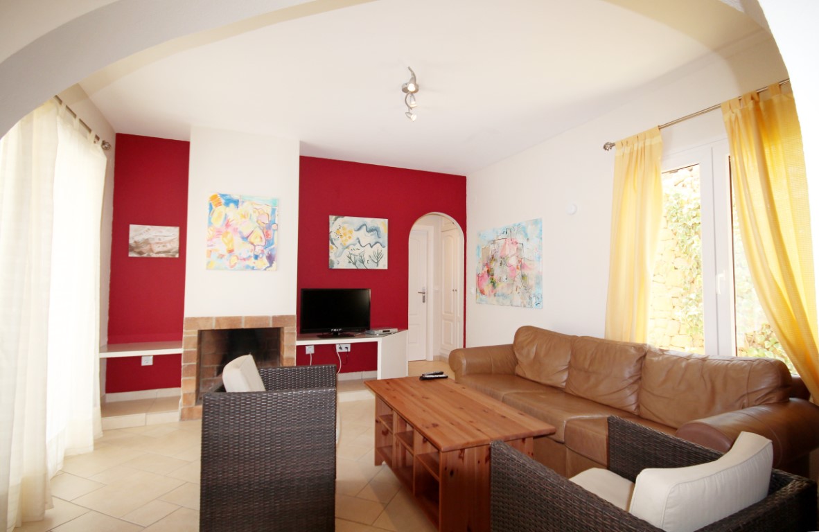 Salon avec mur d'accent rouge, cheminée en briques traditionnelle, canapé en cuir marron et fauteuils en osier sombre, baigné de lumière naturelle latérale.