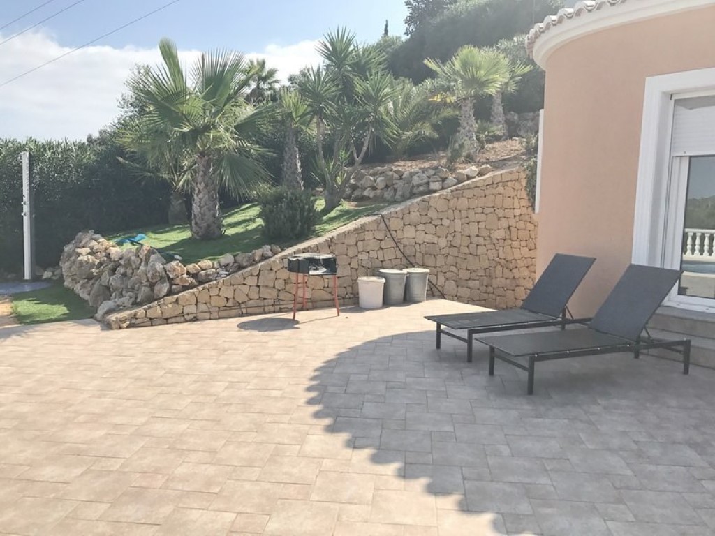 Terrasse dallée avec chaises longues et barbecue, bordée par un mur de soutènement en pierre naturelle et un jardin paysager avec des palmiers.