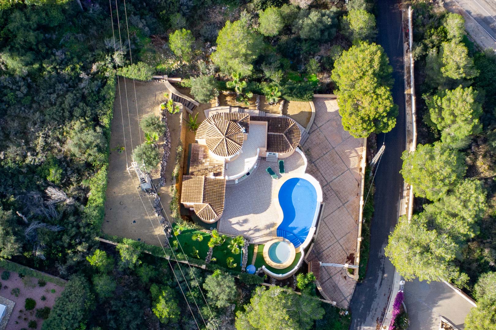 Vue aérienne de la villa présentant ses toits en tuiles, une piscine en forme de haricot et un jacuzzi circulaire, le tout niché dans une forêt méditerréenne dense.