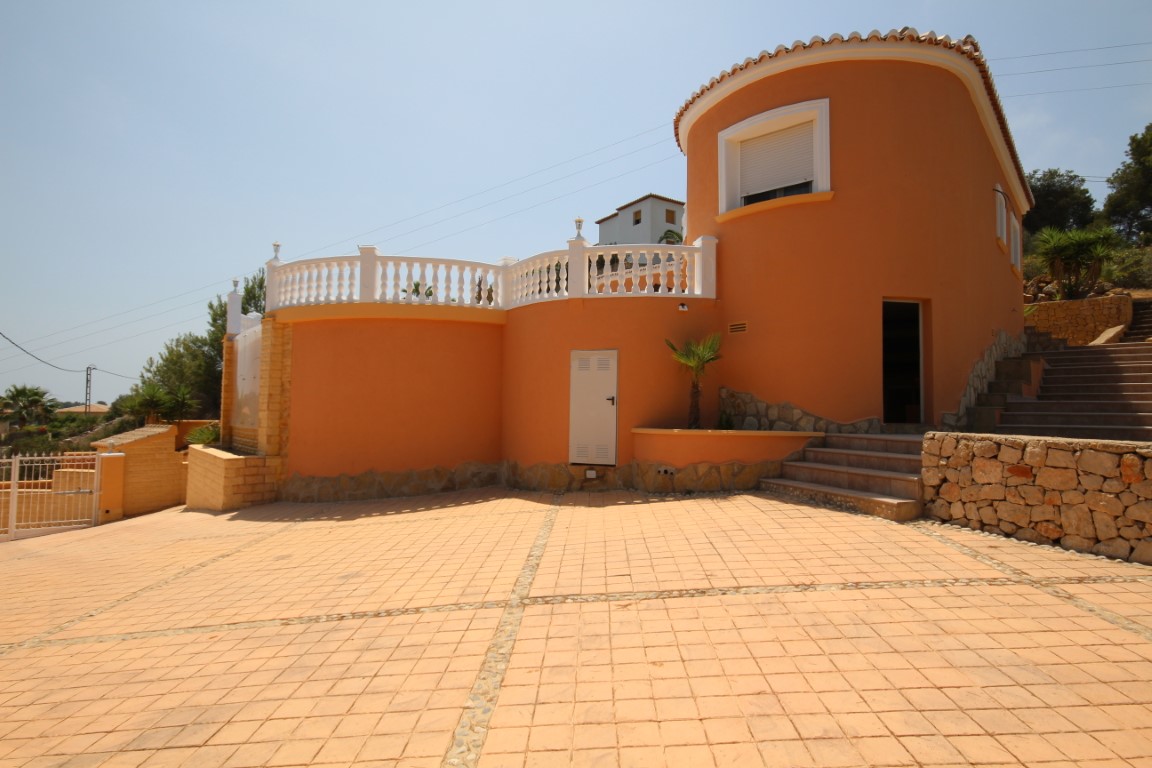 Vue extérieure de la villa avec façade ocre, murs courbes et vaste allée pavée. La terrasse est soulignée par des balustrades blanches et des murs en pierre naturelle.