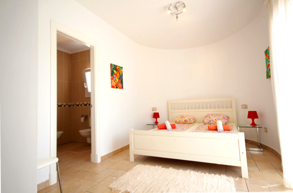 Chambre à coucher avec mur incurvé, cadre de lit en bois blanc et salle de bain attenante carrelée dans des tons naturels.