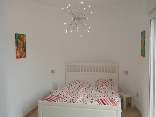 Chambre avec lit en bois blanc, lustre moderne en forme d'étoile et murs incurvés. Comprend un sol en terre cuite et un accès vers l'extérieur.