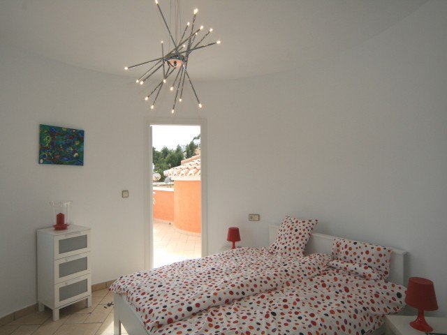 Chambre lumineuse avec mur courbe, lustre design moderne et accès direct à une terrasse privée. Dispose de meubles blancs et d'un sol en carrelage.