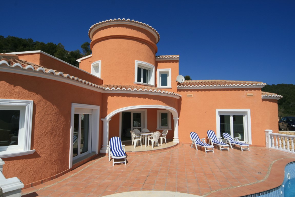 Extérieur de villa méditerranéenne aux tons terre cuite avec tour circulaire, vaste terrasse carrelée, chaises longues rayées et porche voûté ombragé.