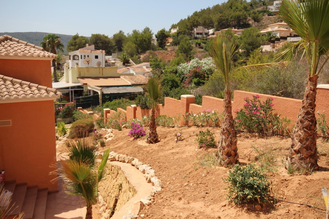 Jardin méditerranéen en terrasses avec de jeunes palmiers, des lauriers-roses et des murs d'enceinte ocre s'ouvrant sur le paysage vallonné environnant.