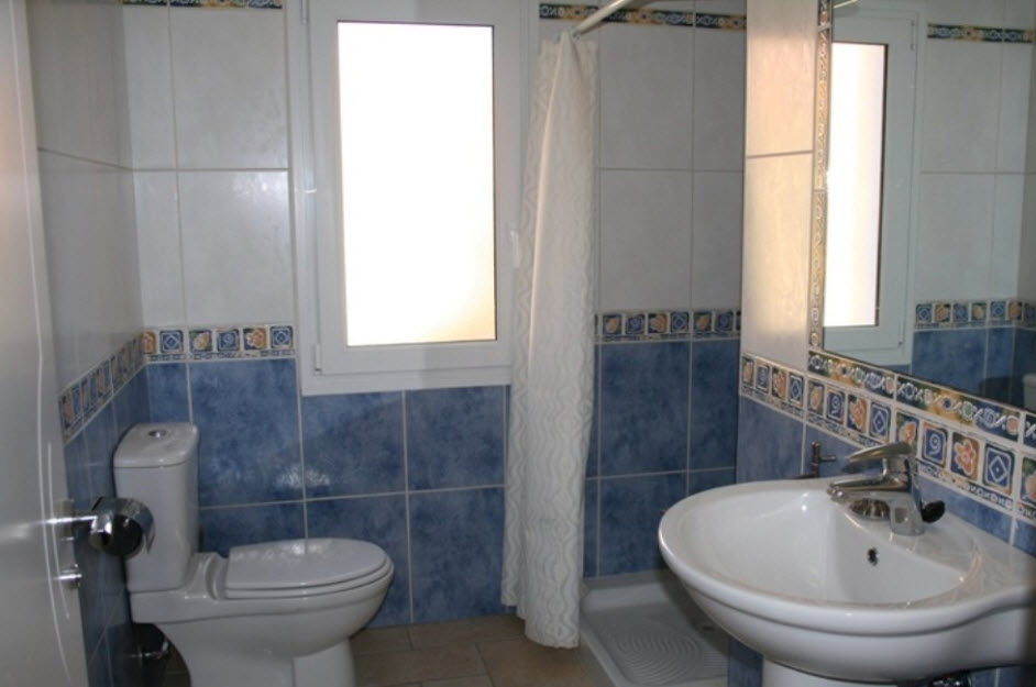 Salle de bain avec carrelage bleu et blanc, lavabo sur colonne, receveur de douche avec rideau et fenêtre en verre dépoli offrant une luminosité naturelle.