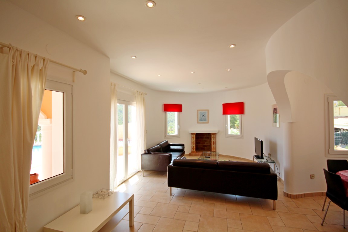 Salon lumineux avec mur incurvé, cheminée intégrée et spots encastrés. Les grandes baies vitrées offrent un accès direct vers l'extérieur.