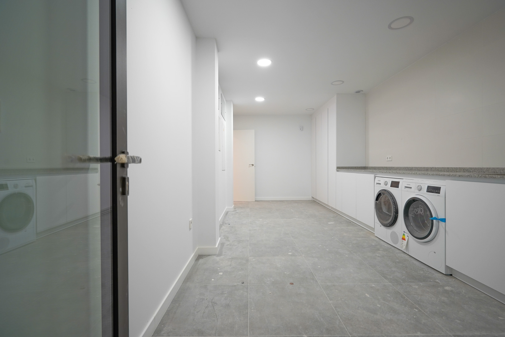 Buanderie spacieuse au rez-de-chaussée avec placards blancs, plan de travail en pierre, appareils Siemens et carrelage gris moderne.