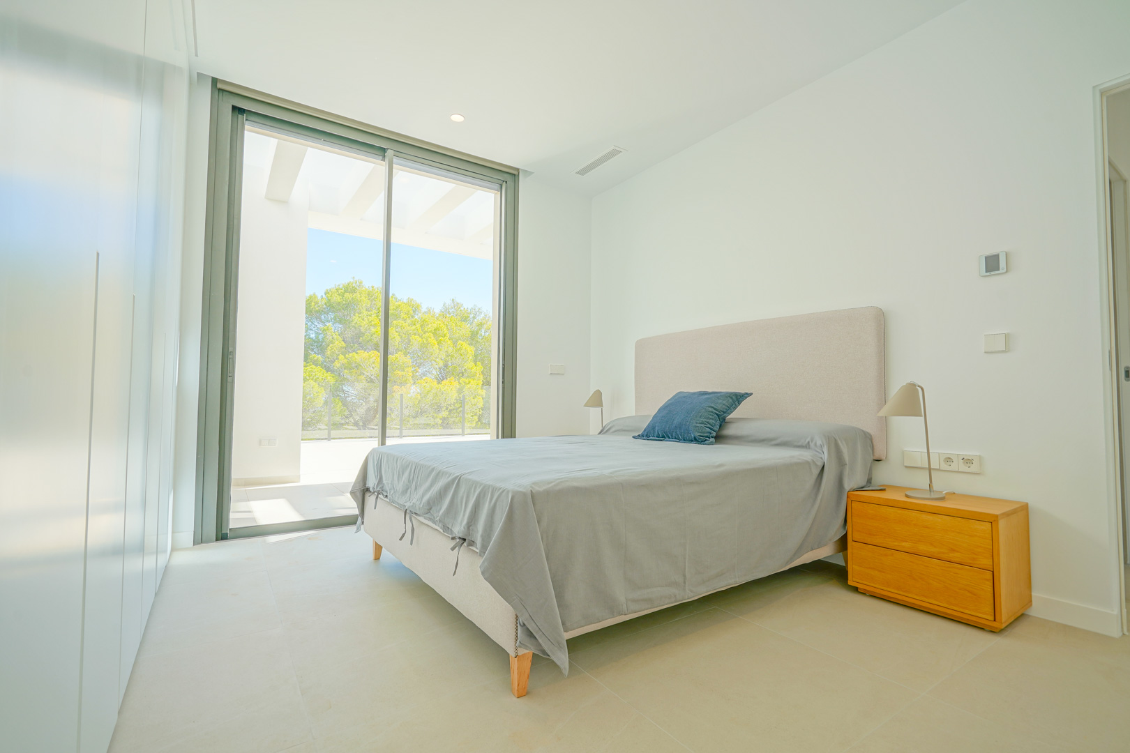 Chambre lumineuse avec baies vitrées, accès direct à la terrasse sous pergola, placards intégrés minimalistes et vue sur la zone boisée de Moraira.