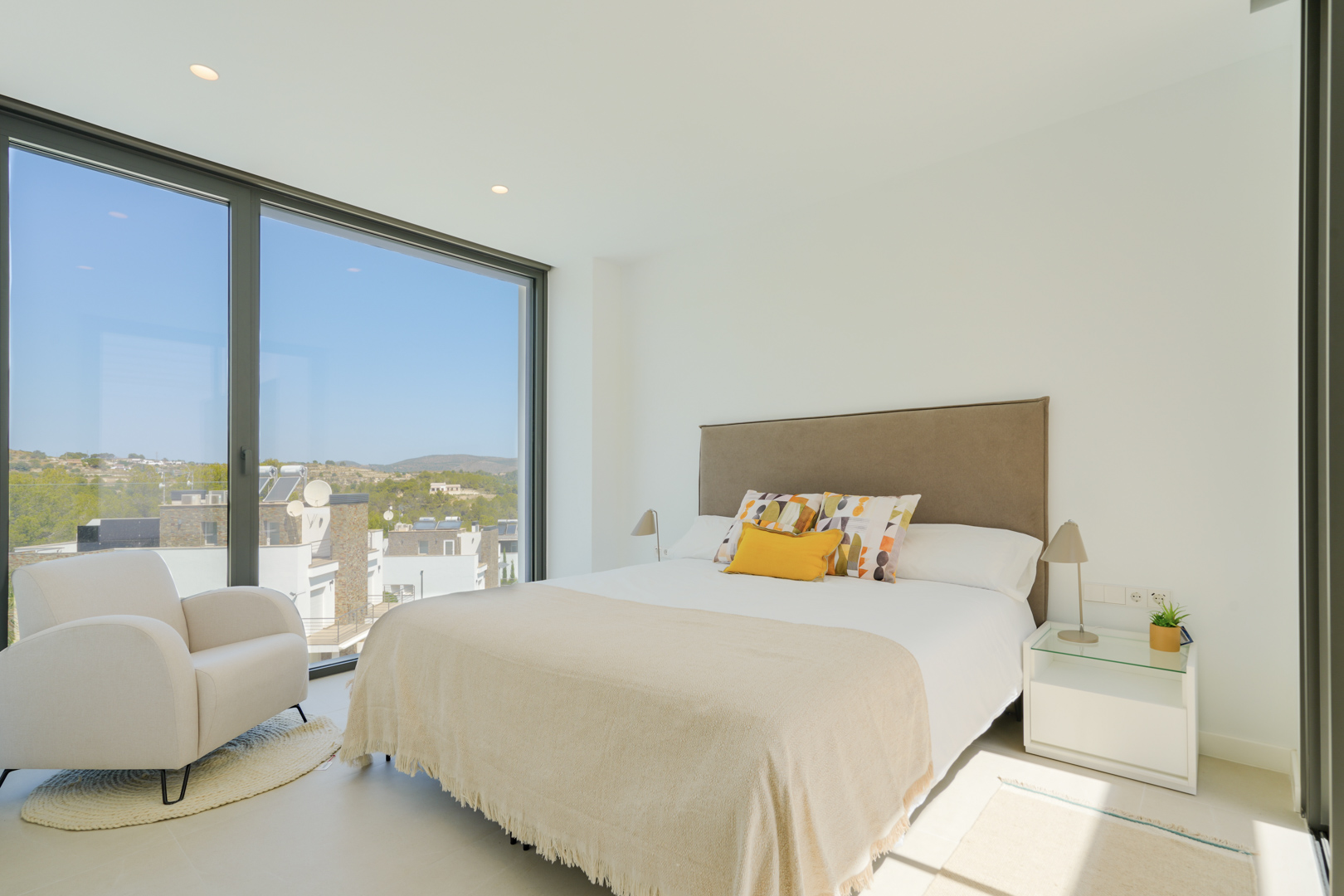 Chambre lumineuse avec de grandes baies vitrées, un lit double à tête de lit rembourrée et un fauteuil design offrant une vue dégagée sur les collines environnantes.