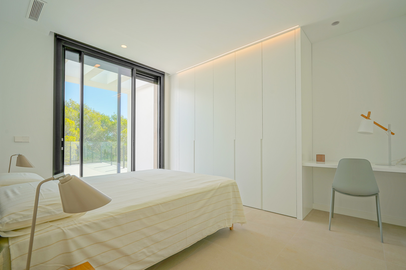 Chambre lumineuse avec placards intégrés blancs, éclairage LED, coin bureau et accès direct à une terrasse offrant une vue sur la zone boisée.
