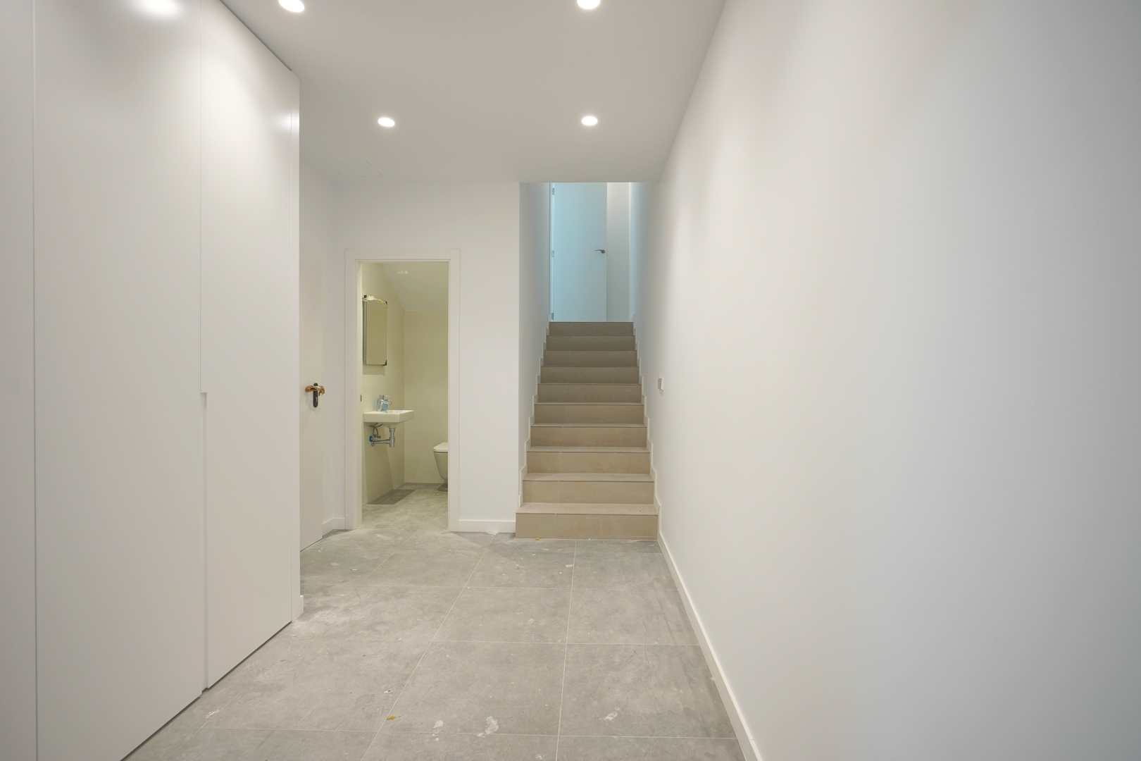 Couloir de distribution avec carrelage gris, placards intégrés blancs et escalier en pierre. Accès visible à un cabinet de toilette et éclairage par spots LED.