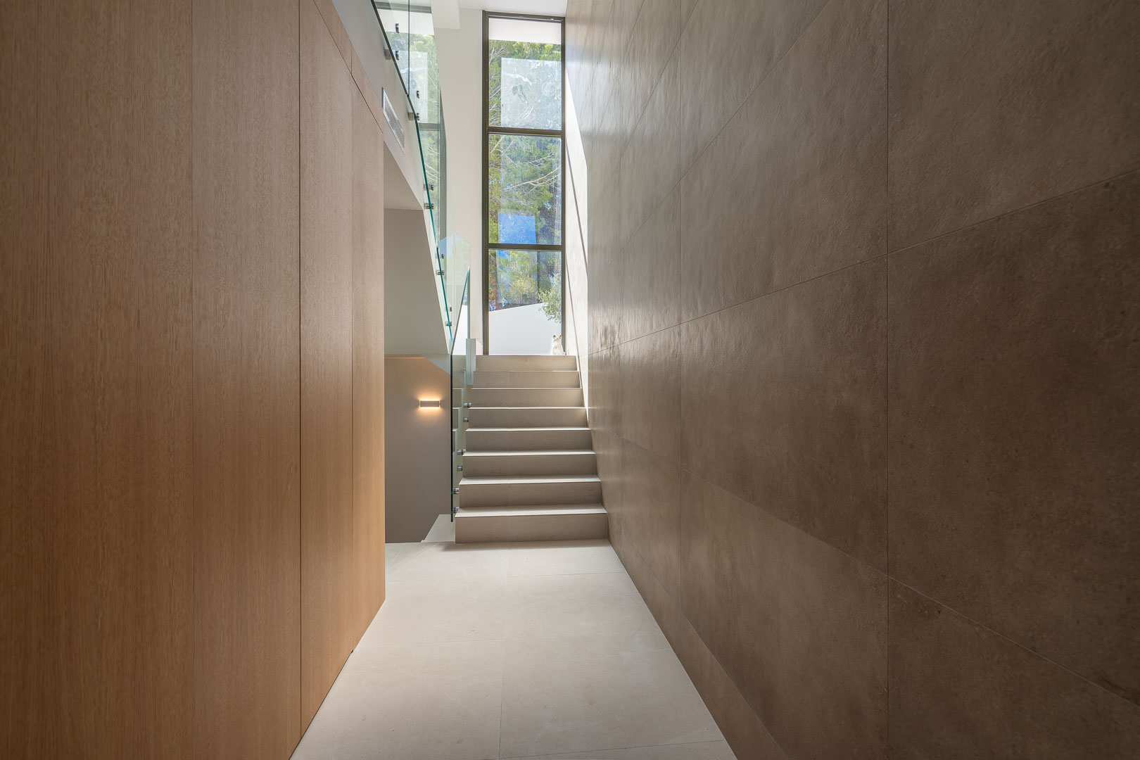 Couloir moderne avec boiseries, murs en pierre grand format et escalier à garde-corps en verre, baigné de lumière par une haute fenêtre verticale.