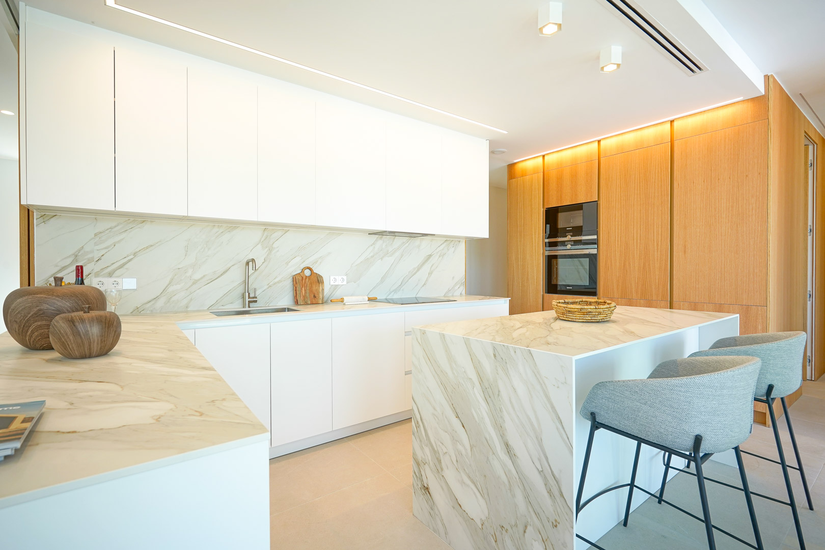 Cuisine moderne avec meubles blancs sans poignées, plans de travail effet marbre et îlot central. Un mur en bois intègre l'électroménager sous un éclairage LED encastré.