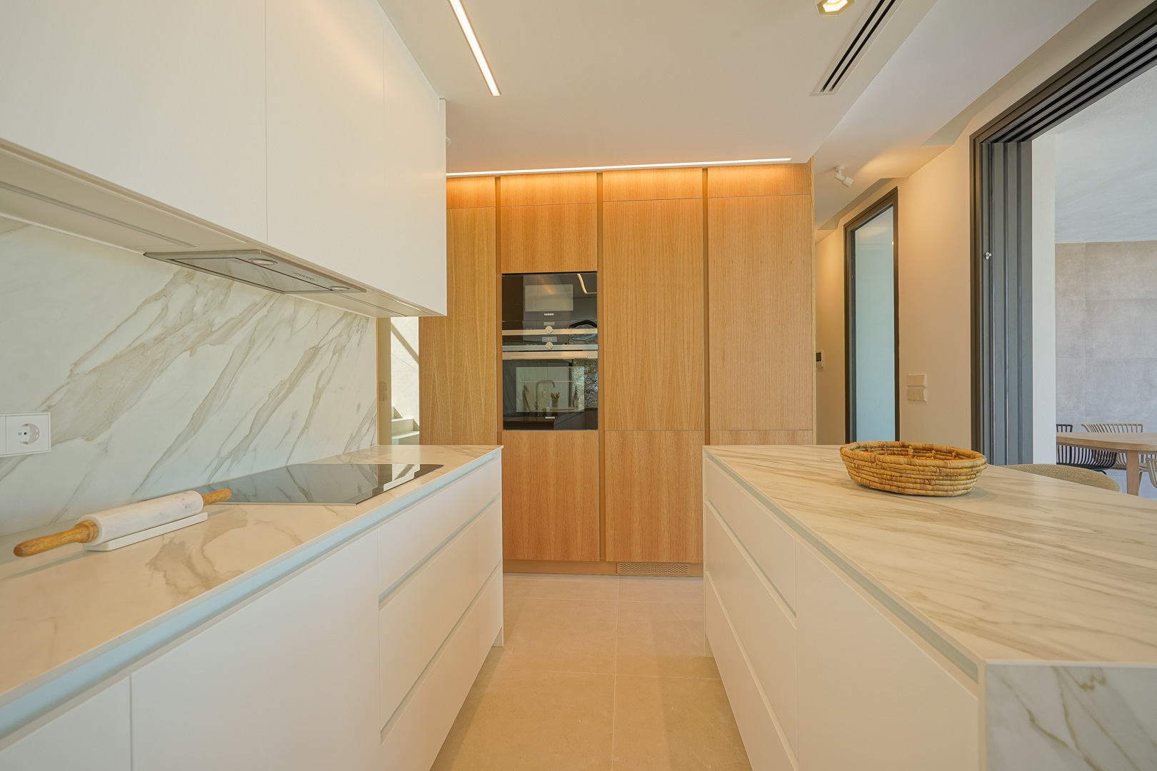 Cuisine moderne avec plans de travail en marbre veiné, placards blancs minimalistes et un mur de rangement en bois avec électroménager encastré et éclairage linéaire.