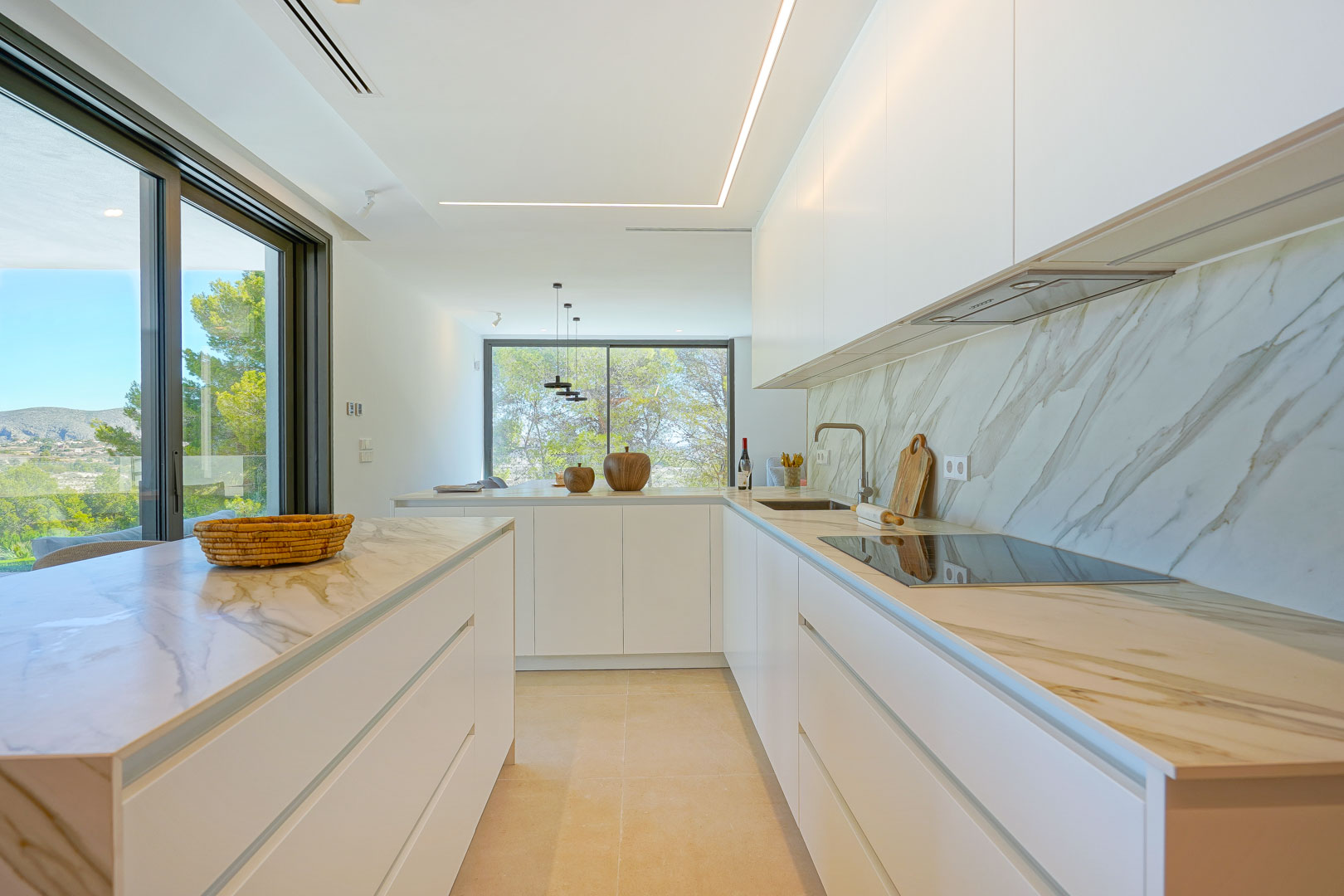 Cuisine ouverte minimaliste avec plans de travail effet marbre, meubles blancs sans poignées et éclairage LED linéaire. Les baies vitrées offrent une vue sur la zone boisée.