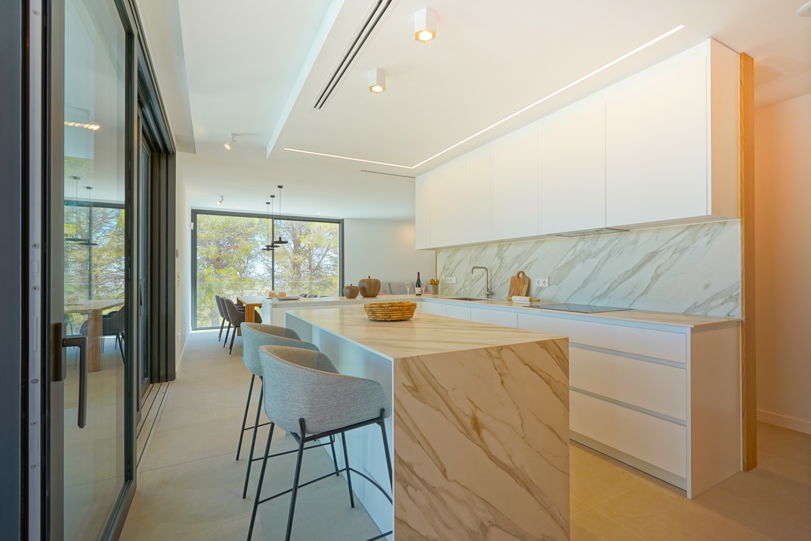 Cuisine ouverte moderne avec îlot en cascade effet marbre et placards blancs épurés. Éclairage LED intégré et accès direct à la terrasse par de larges baies.