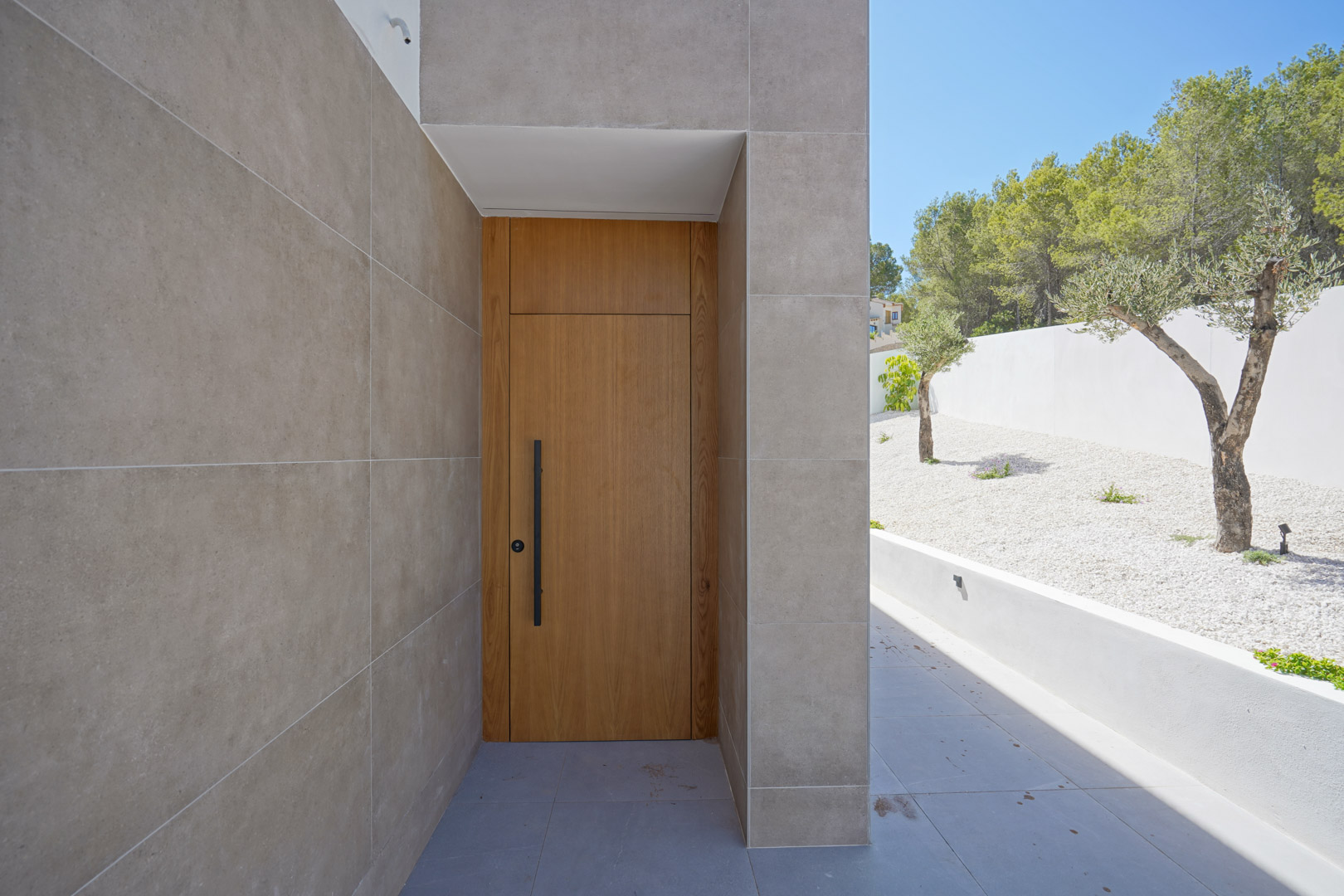 Entrée principale avec porte en bois massif et poignée noire, encadrée par une façade en grès cérame et un jardin méditerranéen de gravier blanc.