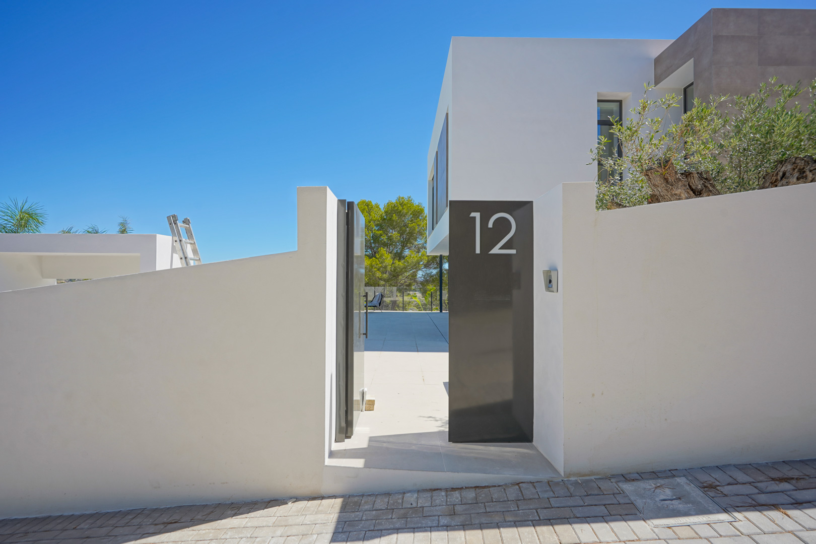 Entrée principale moderne avec mur d'enceinte blanc, portillon métallique gris anthracite affichant le numéro 12 et accès à un patio dallé sous un ciel azur.