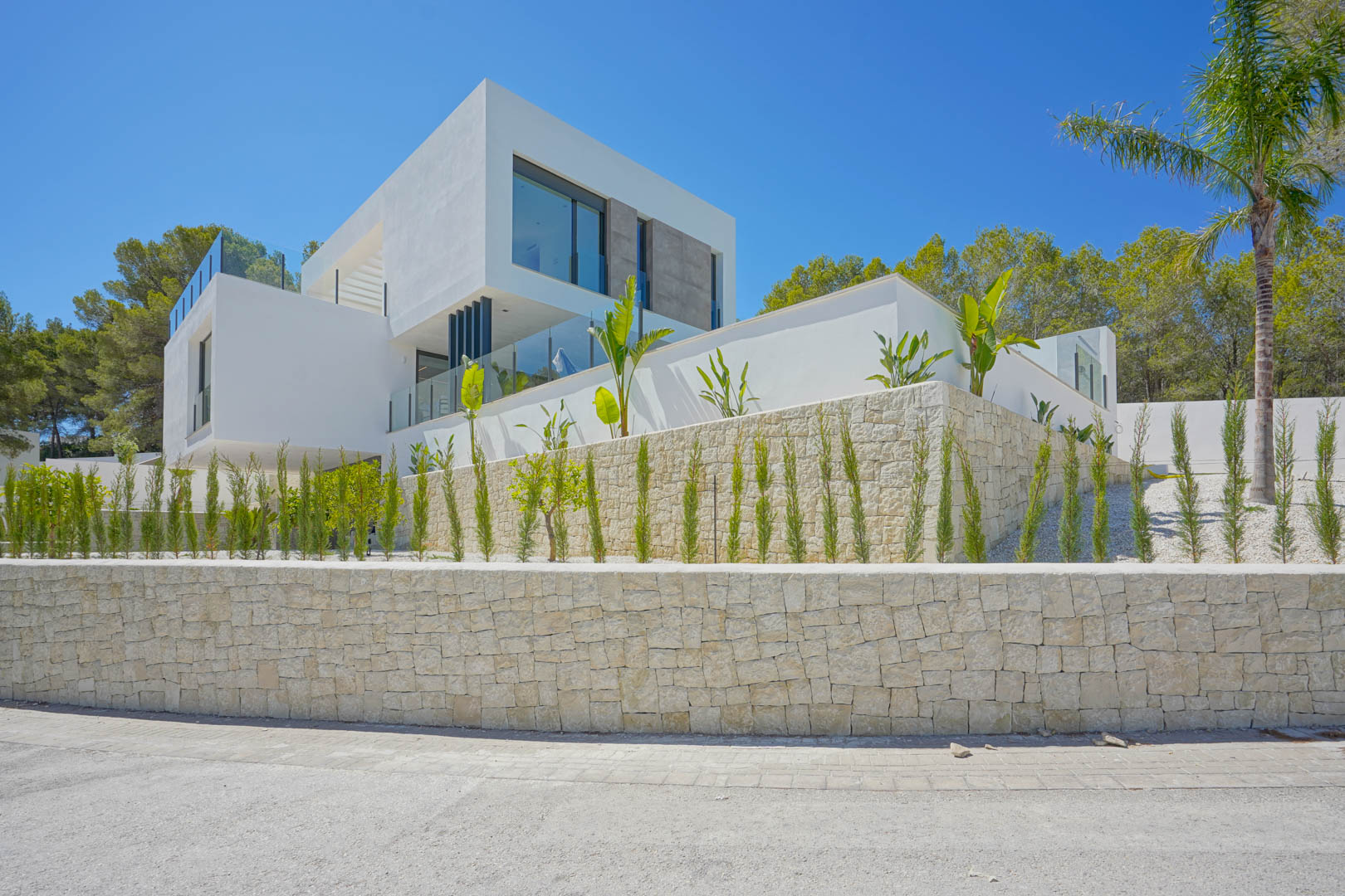 Façade contemporaine aux volumes blancs épurés, agrémentée de murs en pierre sèche, de larges baies vitrées et d'un jardin méditerranéen avec cyprès et palmiers.