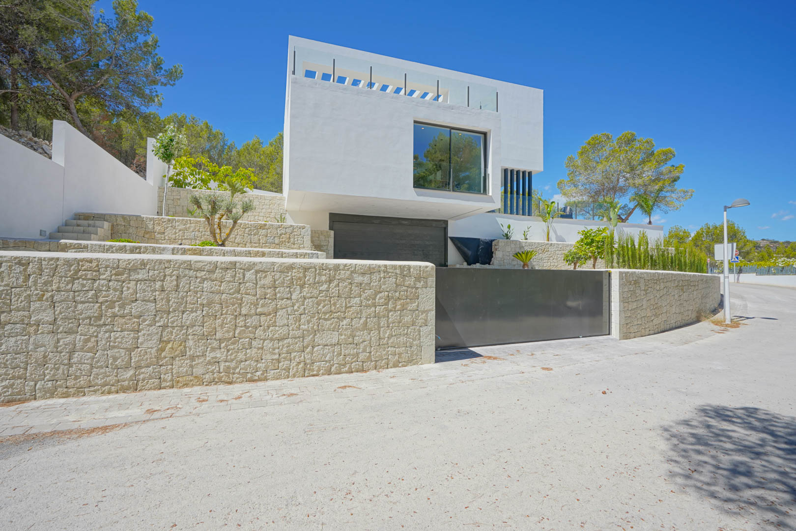 Façade extérieure moderne avec murs de soutènement en pierre naturelle, entrée de garage double et jardin méditerranéen en terrasses.