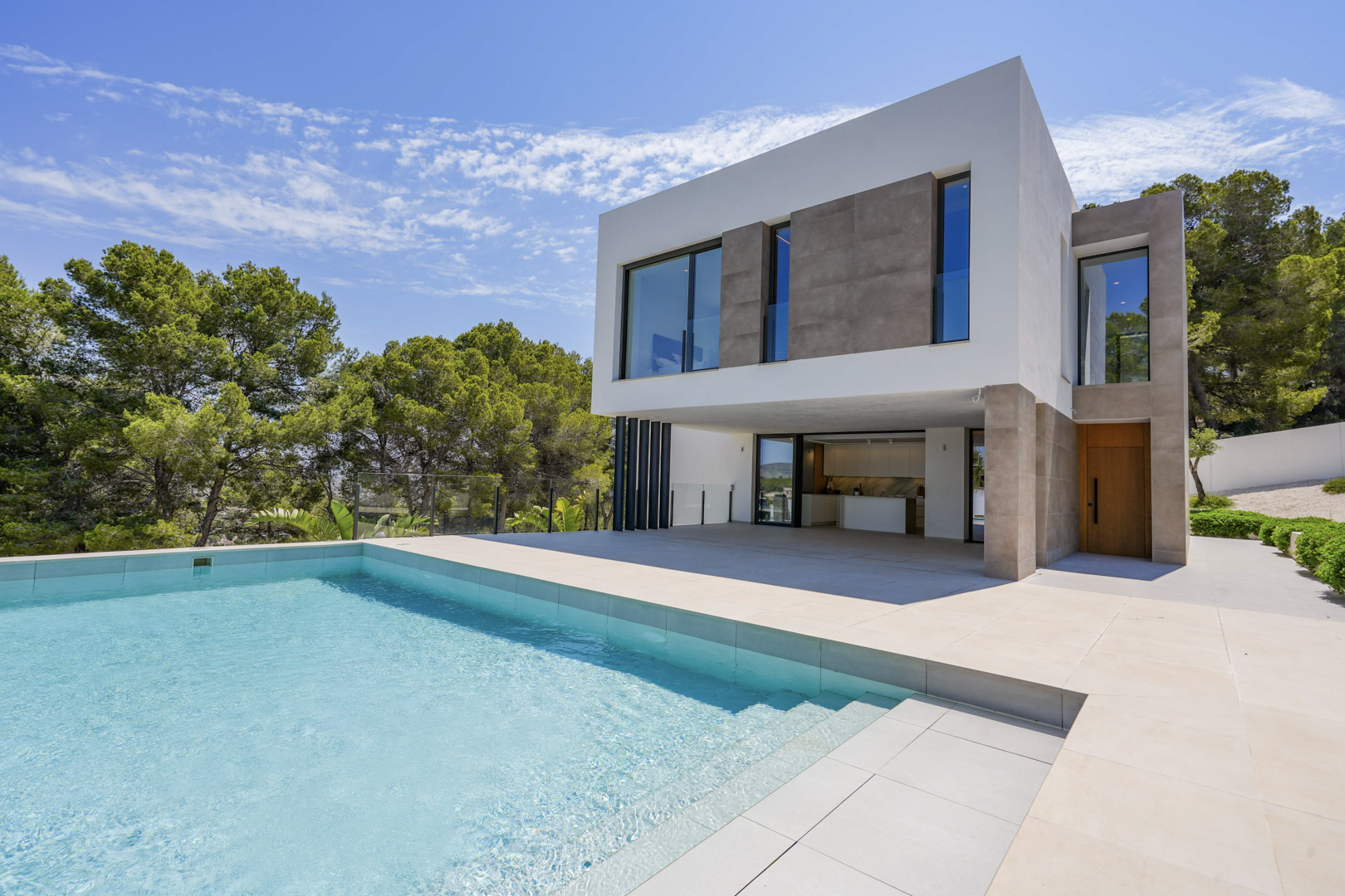 Façade moderne de la villa avec piscine à débordement et vaste terrasse en pierre, nichée dans un environnement boisé sous un ciel dégagé.