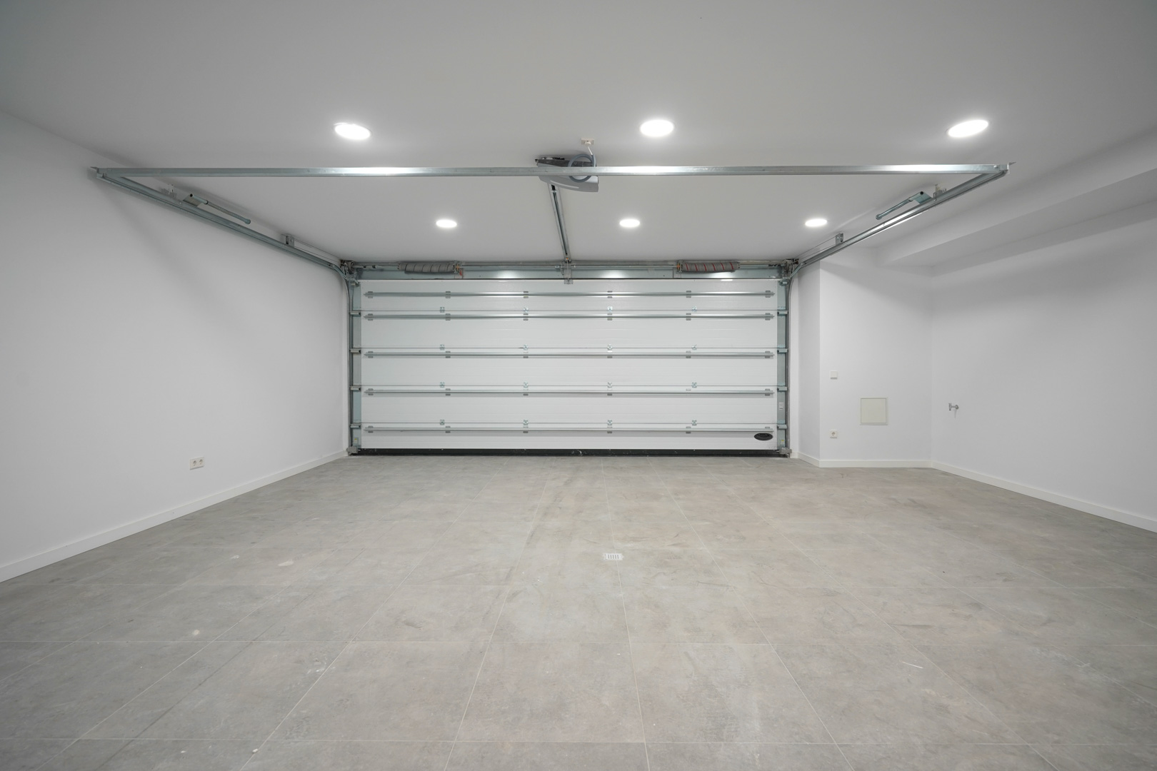 Grand garage pour deux voitures avec porte sectionnelle automatique, sol en grès gris et éclairage LED encastré au plafond.