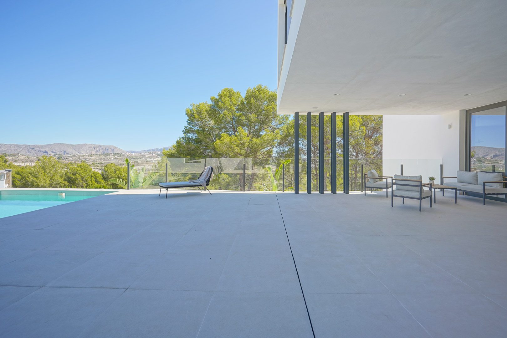 Grande terrasse avec carrelage grand format, salon d'été abrité et garde-corps en verre offrant une vue panoramique sur la pinède environnante.