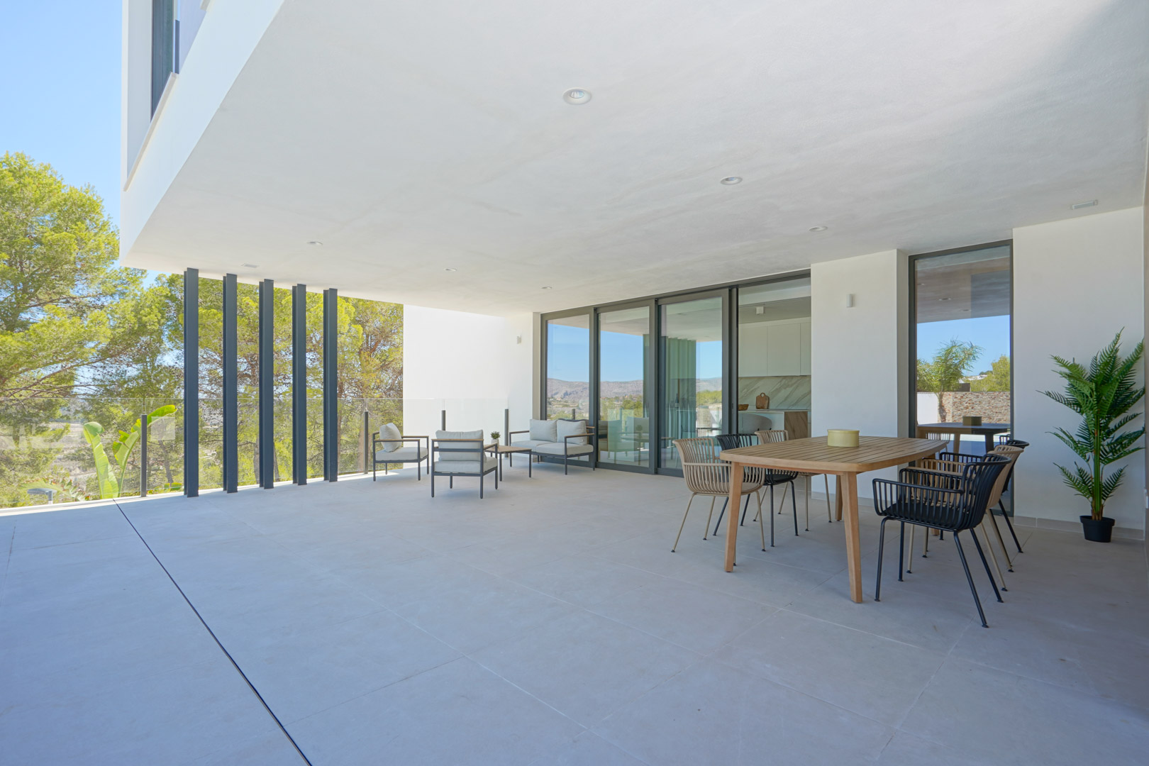 Grande terrasse couverte avec coin repas et salon d'été, dotée de garde-corps en verre et offrant une vue dégagée sur la zone boisée environnante.