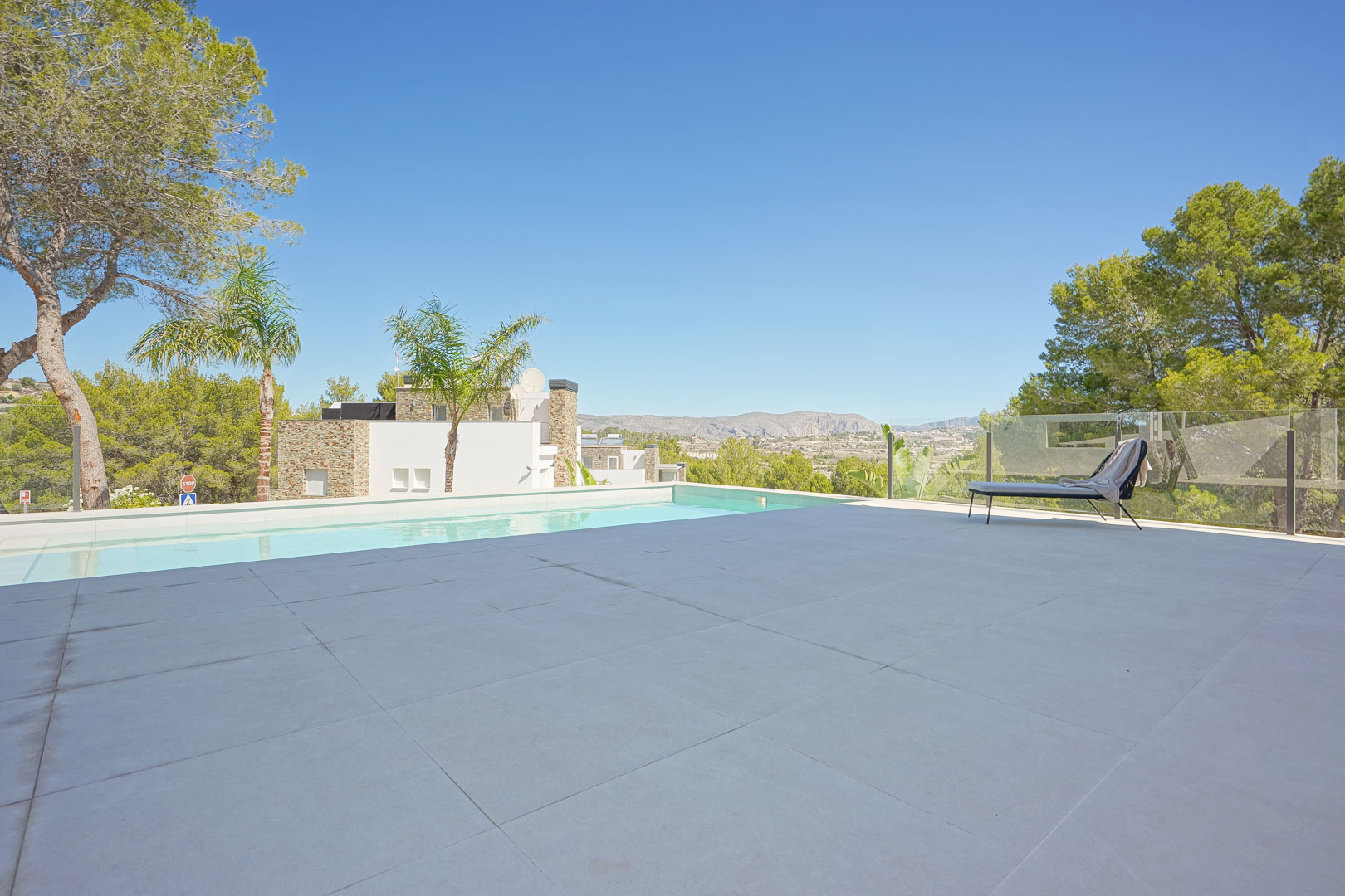 Grande terrasse en carrelage clair avec piscine à débordement, garde-corps en verre et vue panoramique sur la vallée et la pinède environnante.