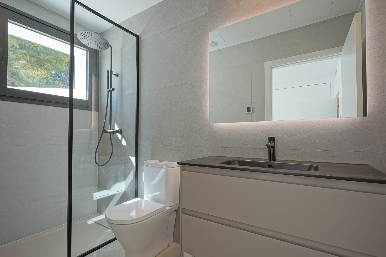 Salle de bain moderne avec carrelage gris, douche à l'italienne avec cadre noir et miroir rétroéclairé. Elle dispose d'un meuble vasque avec plan de travail sombre et épuré.