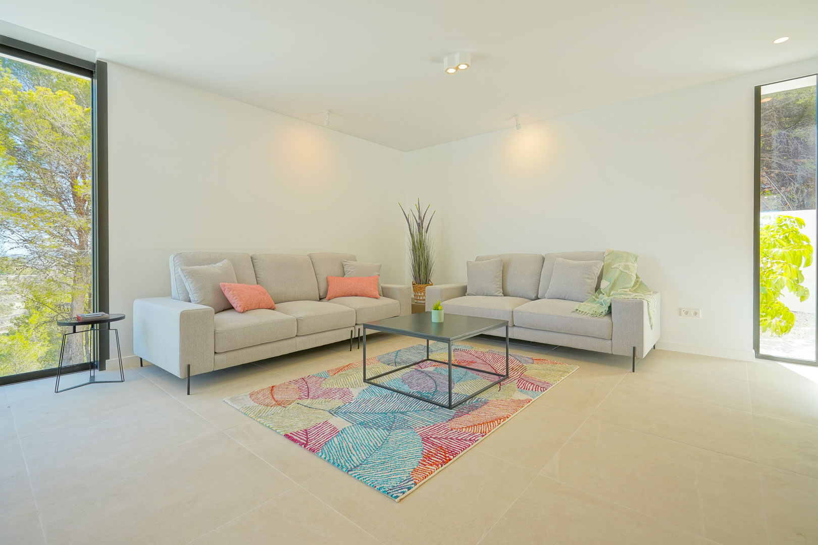 Salon lumineux avec canapés gris, tapis aux motifs colorés et grandes baies vitrées offrant une vue sur la pinède environnante.