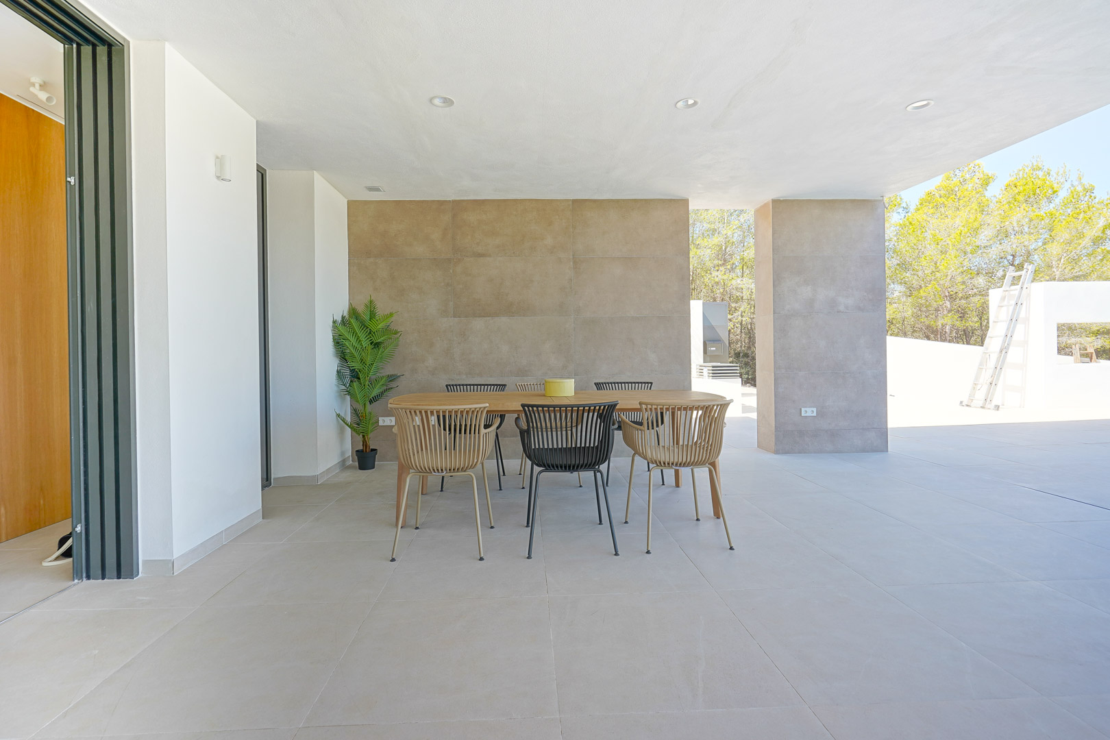 Terrasse couverte avec coin repas extérieur, comprenant une table en bois, un mur d'accent texturé et un sol en dalles grand format avec vue sur la pinède.