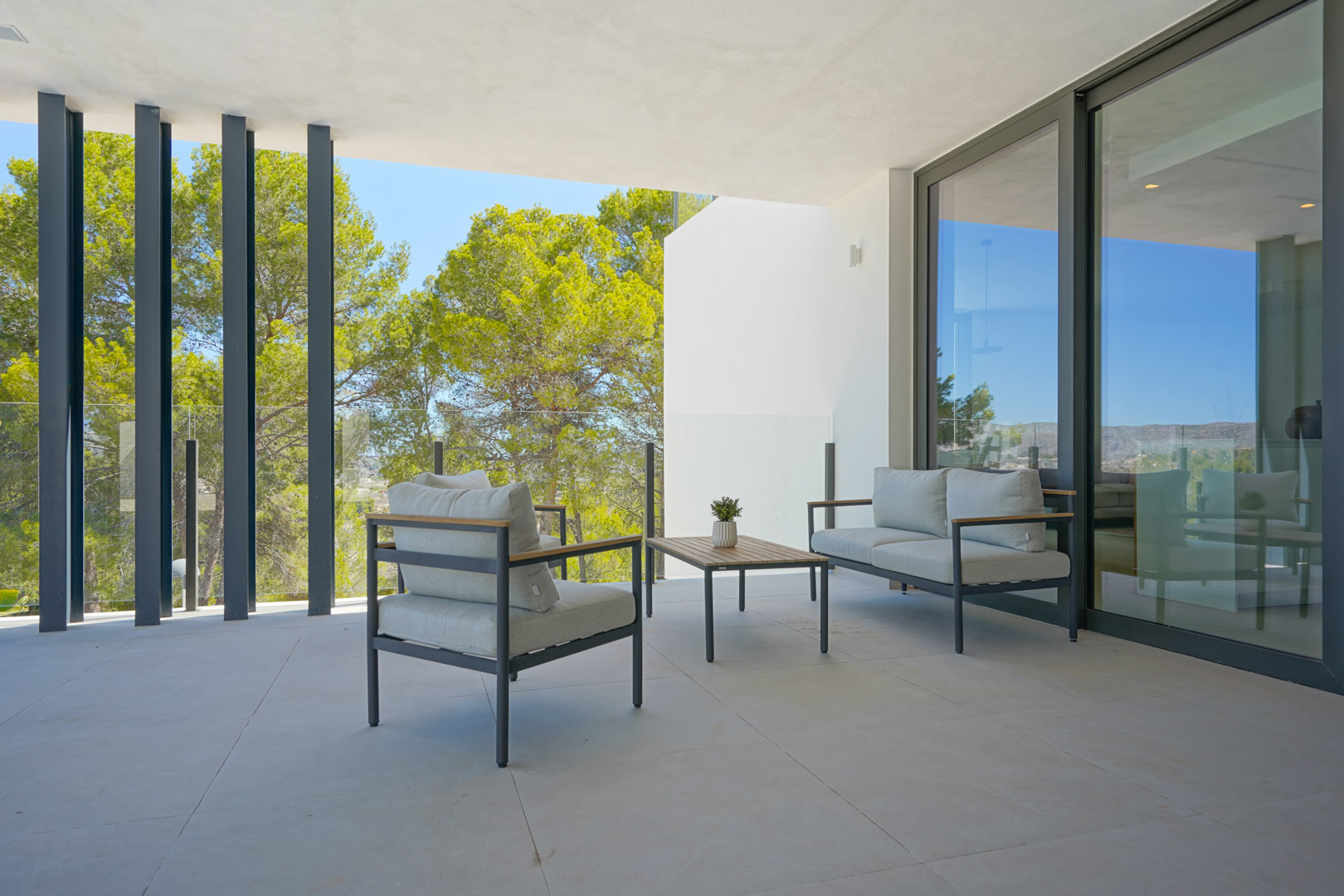 Terrasse couverte avec mobilier de salon moderne, garde-corps en verre et vue sur la pinède à travers des éléments architecturaux verticaux.