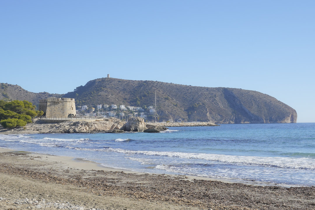 Vue de la plage de Moraira avec sa tour de guet historique en pierre et le promontoire du Cap d'Or bordant les eaux turquoise.