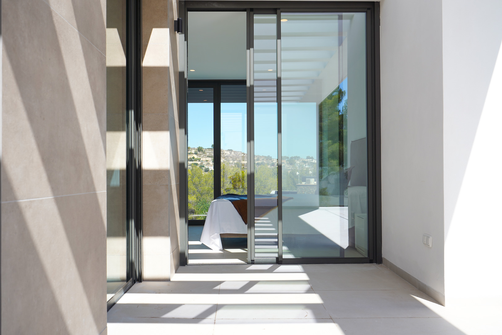 Vue de la terrasse avec revêtement en pierre et baies vitrées coulissantes ouvrant sur une chambre. Ombres linéaires de la pergola sur le sol carrelé.