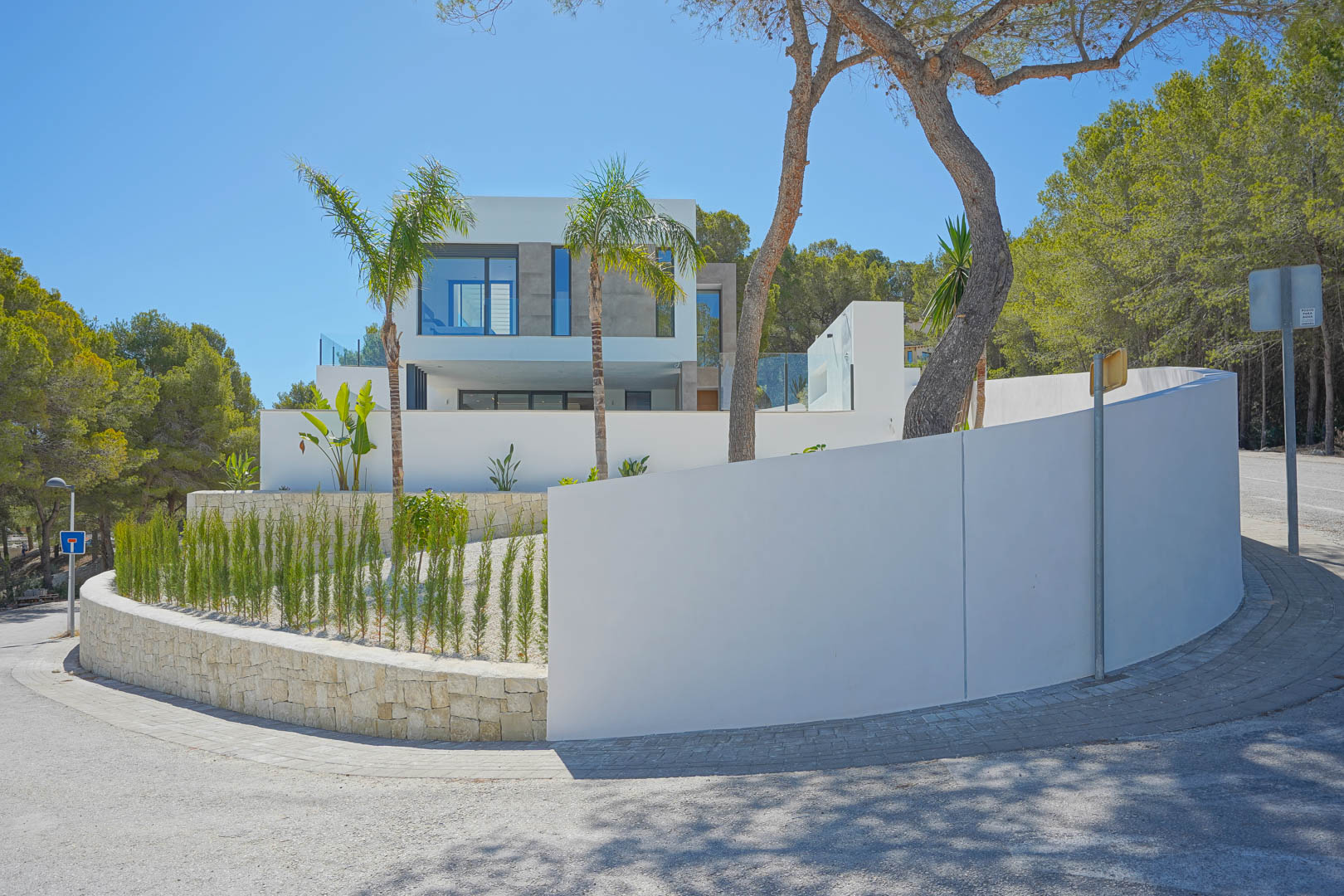 Vue extérieure de la villa moderne avec façades blanches, murs incurvés et jardin méditerranéen composé de cyprès et de palmiers.