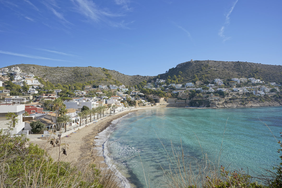 Vue panoramique de la plage d'El Portet à Moraira, avec ses eaux turquoise, sa promenade bordée de palmiers et les collines résidentielles environnantes.