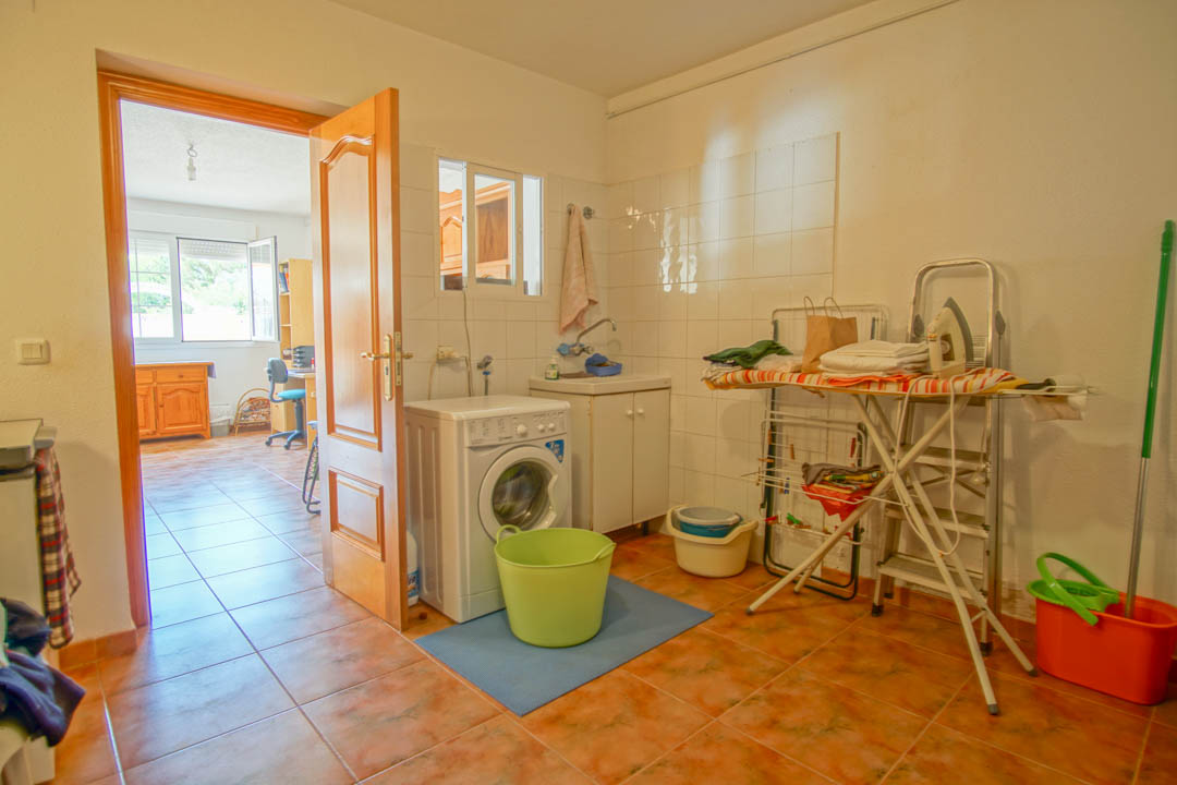 Buanderie fonctionnelle avec lave-linge, évier et espace de repassage, dotée d'un sol en terre cuite et d'une porte en bois massif menant au bureau.