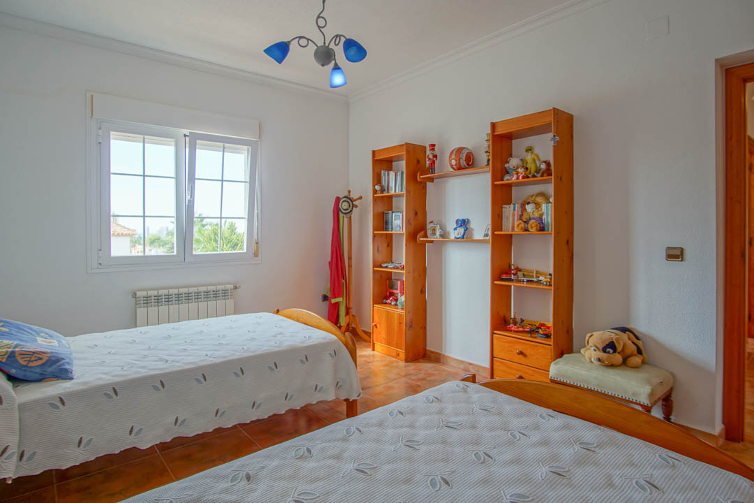 Chambre avec deux lits simples, étagères en bois massif et sol en céramique. Comprend un radiateur mural et une fenêtre à double vitrage apportant une luminosité naturelle.
