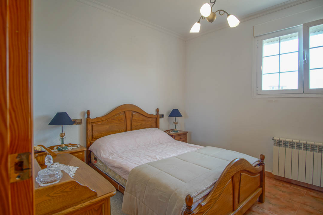 Chambre avec mobilier en bois massif, sol en grès et radiateur. La pièce comprend un cadre de lit et des tables de chevet assortis, éclairés par une fenêtre à croisillons.