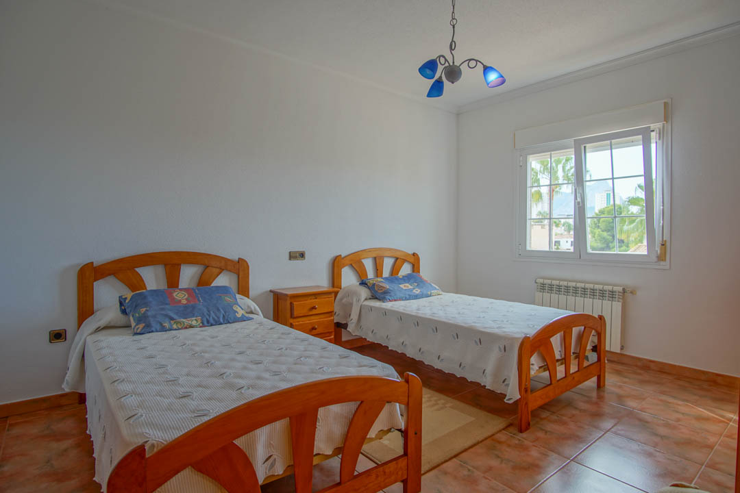 Chambre lumineuse avec deux lits simples en bois massif, sol en céramique et fenêtre offrant une vue sur les palmiers et les montagnes environnantes.